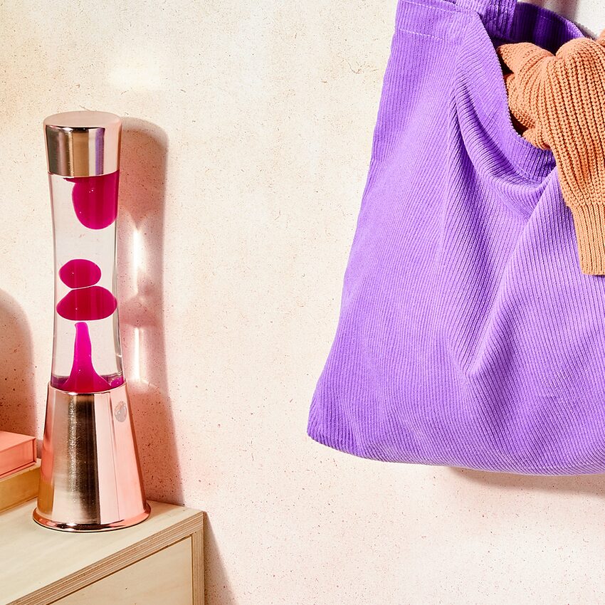 Lampe à lave rose posée sur un meuble, à côté d'un tote bag violet en velours côtelé