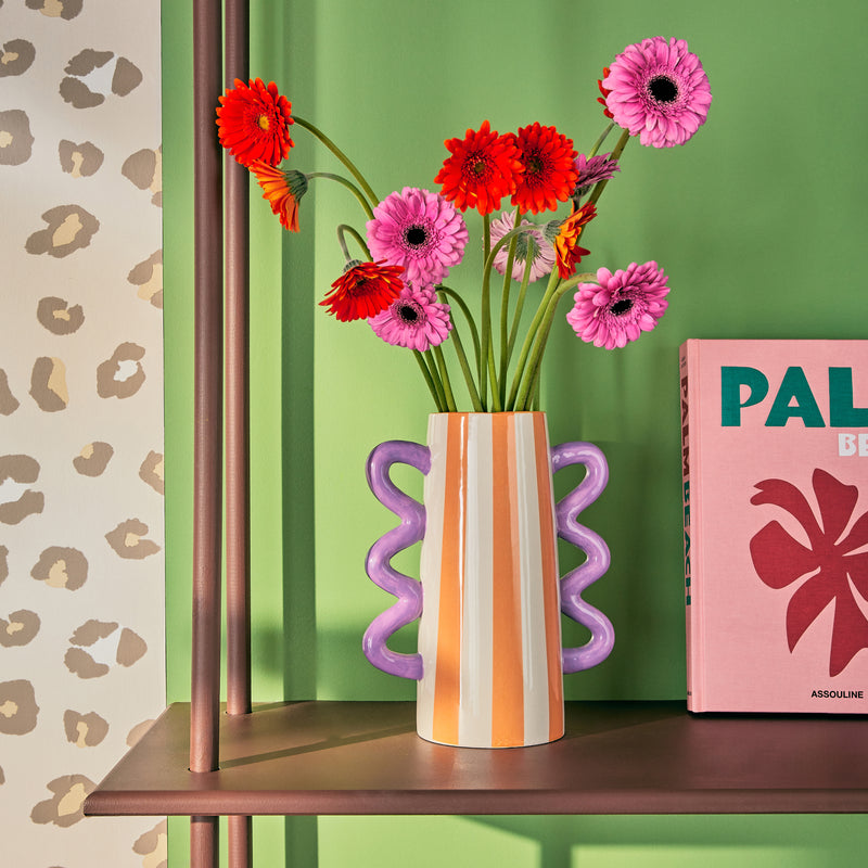 Vase rayé blanc et orange à anses ondulées violettes, mur vert franc et papier peint léopard