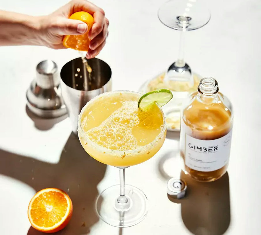 Verre à cocktail rempli d'une boisson apéritive sans alcool au gingembre, bouteille de concentré de gingembre bio de la marque Gimber, main pressant une orange