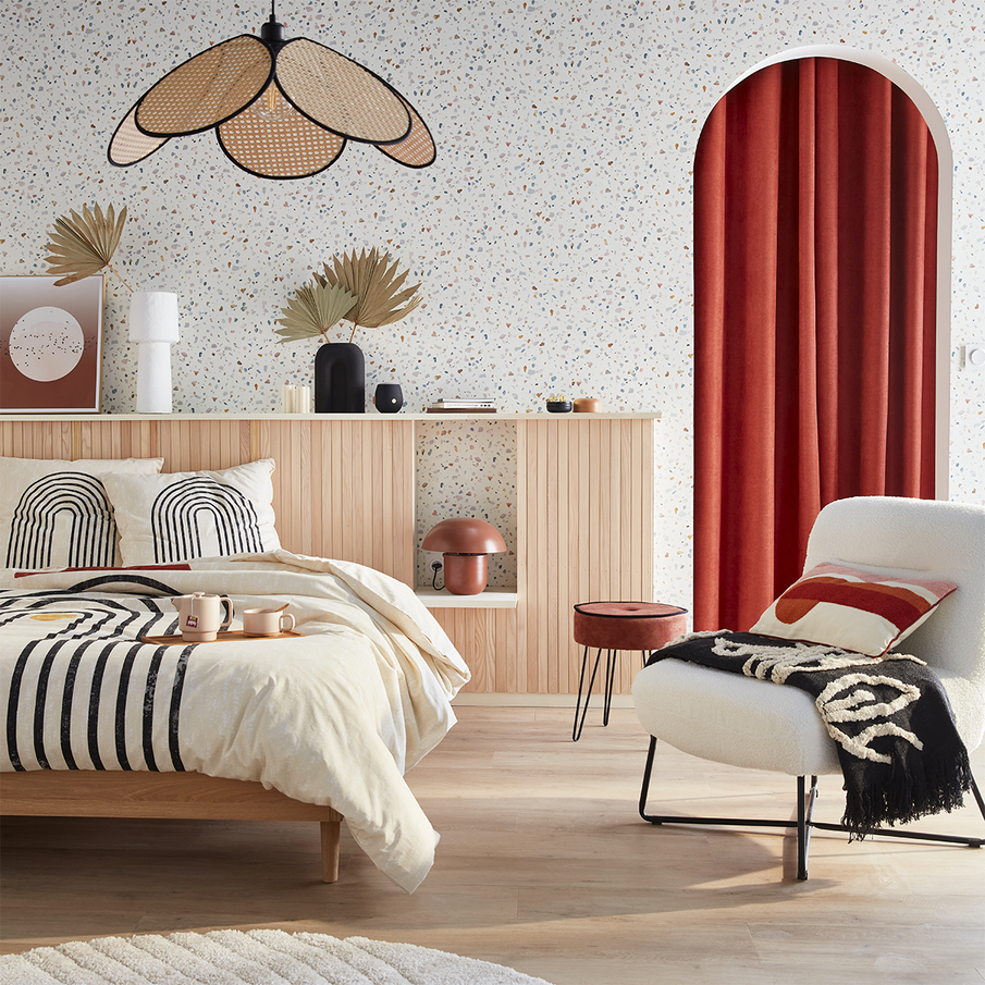 Chambre avec lit en bois clair, linge de lit beige à motifs graphiques noirs, fauteuil blanc avec plaid et coussin rouge, mur en terrazzo clair, tête de lit en lattes de bois et suspension en rotin en forme de pétales