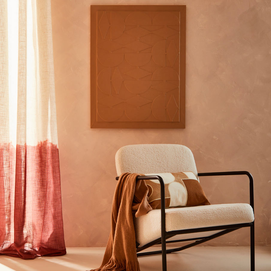 Fauteuil en tissu bouclette et structure métallique, sur lequel sont posés un coussin et un plaid, devant un mur couleur beige et un tableau brun aux motifs géométriques
