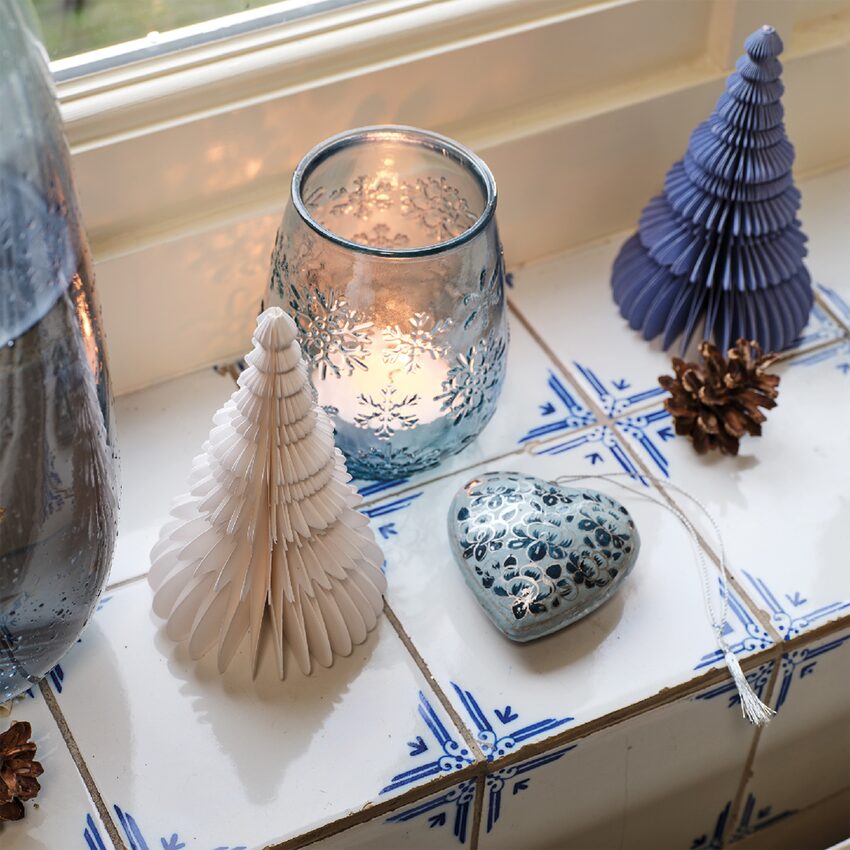 Photophore avec bougie allumée et deux sapins en papier posés sur un rebord de fenêtre