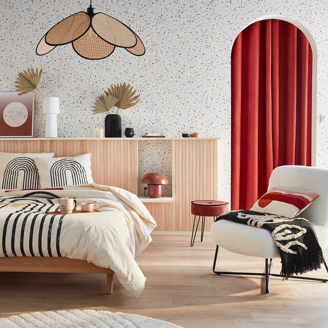 Chambre avec lit en bois clair, linge de lit beige à motifs graphiques noirs, fauteuil blanc avec plaid et coussin rouge, mur en terrazzo clair, tête de lit en lattes de bois et suspension en rotin en forme de pétales