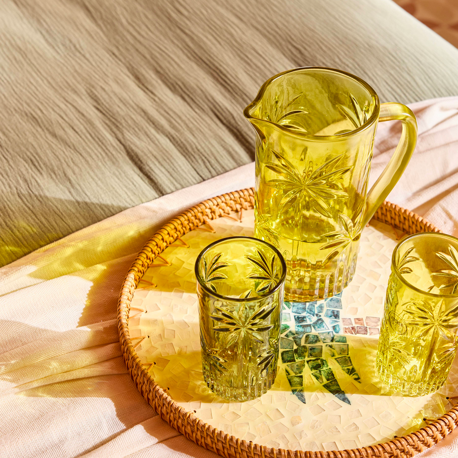 Vaisselle en verre, pichet et verres posés sur un plateau au motif de palmier en mosaïque, posé sur un lit